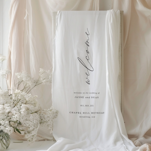 fabric welcome sign for a wedding event, white linen fabric with white text that says "welcome to the wedding of" and then the couples name, wedding date and venue.